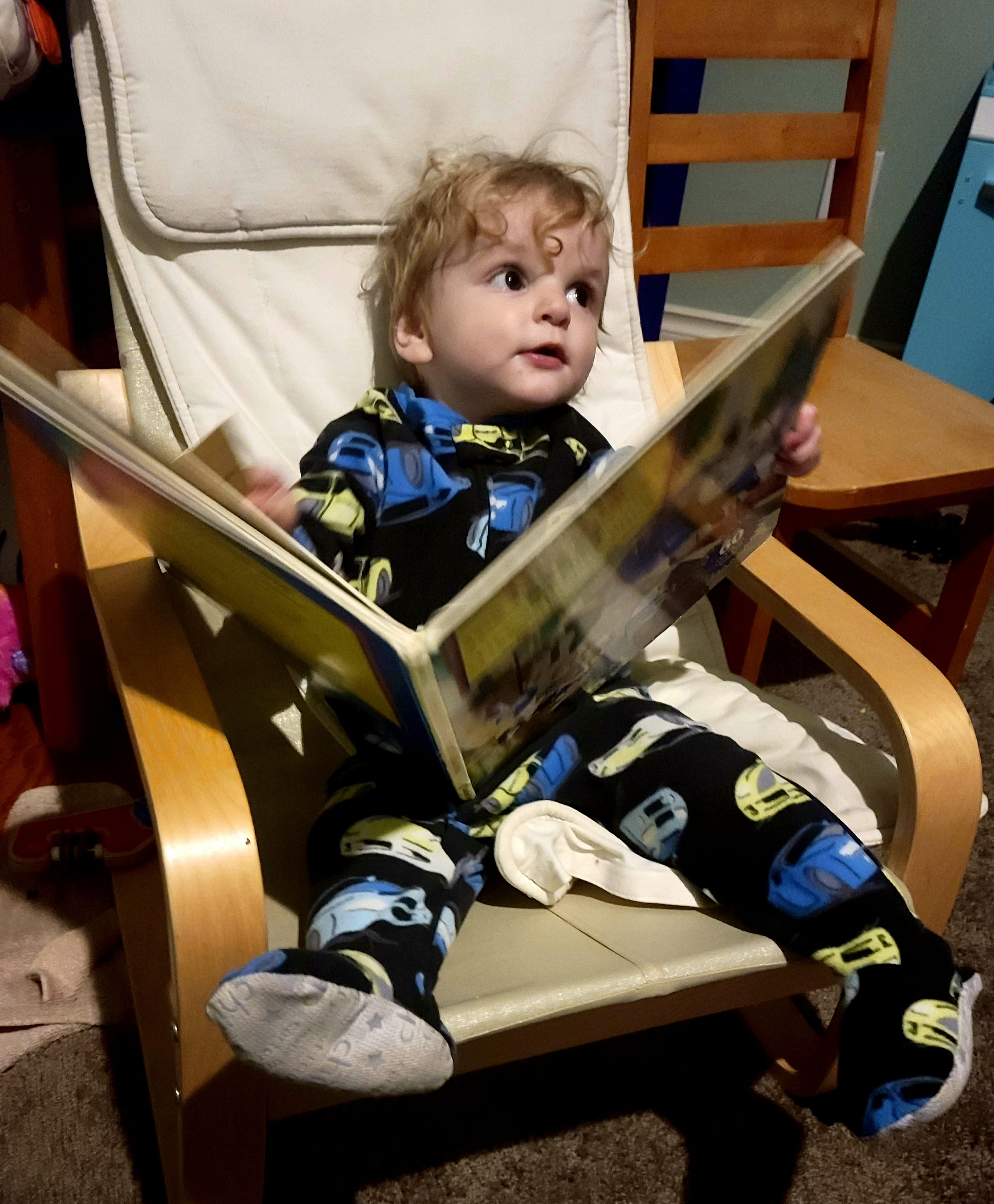 Photo of two-year old boy in footie pajamas sitting in a kid-sized chair with an open children's book on his lap.